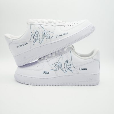 Zwei gestapelte weiße Nike Air Force 1 Sneaker mit blauem 'Pinky Promise' Finger-Motiv, blauen Namen 'Mia' und 'Liam' sowie Datumsangaben '24.02.2026' und '27.01.2021'.