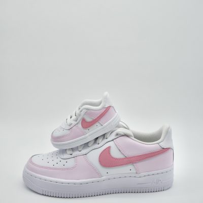 Weißer Babysneaker im hellrosa Akzenten, dunklerem rosa Swoosh und kleinem rosa Detail auf dem Mittelfeld vor neutralem Studio-Hintergrund.