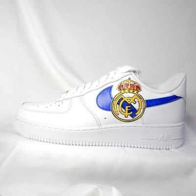 Weiße Nike Air Force 1 Sneakers mit blauen Akzenten und Real Madrid Logo auf der Seite.