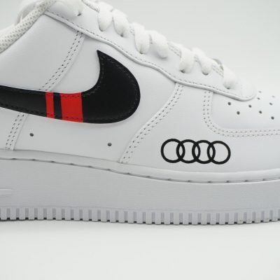 Nahaufnahme eines weißen Nike Air Force 1 mit schwarzem Swoosh, rot-schwarzem Audi Streifen-Motiv und schwarzen Audi-Ringen am Mittelfeld als Auto-Branding.