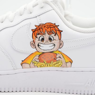 Nahaufnahme eines weißen Nike Air Force 1 mit handgemaltem rothaarigen Jungen, der lachend einen Hamburger und Pommes hält, als verspieltes Comic-Detail an der Seite.