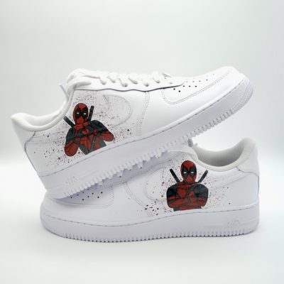 Zwei gestapelte weiße Nike Air Force 1 mit handgemaltem Deadpool-Charakter in roter Comic-Pose und dezenten roten und schwarzen Farbspritzern auf den Seiten.