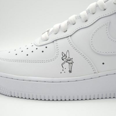 Nahaufnahme eines weißen Nike Air Force 1 mit fein gezeichneter schwarzer Tinker Bell Silhouette aus 'Peter Pan' und kleinen Feenstaub-Sternchen an der Seite.