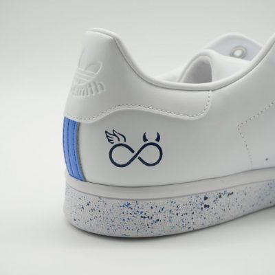Detail-Nahaufnahme der Fersenkappe eines weißen Adidas Stan Smith Custom Sneaker mit Engel- und Teufel-Unendlichkeitssymbol in Blau, blaue Farbspritzer auf der Sohle
