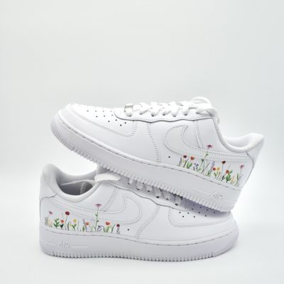 Zwei gestapelte weiße Nike Air Force 1 mit handgemaltem bunten Frühlingsblumen-Streifen entlang der Sohlenkante mit Tulpen, Mohn und Glockenblumen.