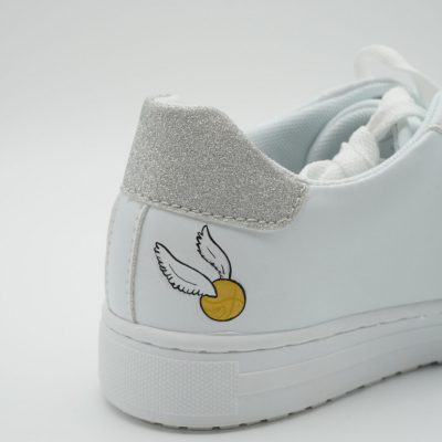 Nahaufnahme eines weißen Sneakers mit handgemaltem goldenen Schnatz mit silbern glitzernden Flügeln an der Seite und glitzernder silberner Fersenkappe als Harry Potter Detail.