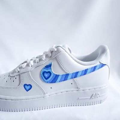 Weißer Nike Air Force 1 Custom Sneaker mit blauem Herz-Design, blaue Streifen-Herzen auf dem Swoosh und kleinem Herz am Mittelfußbereich vor weißem Stoff
