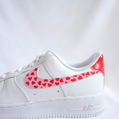 Weißer Nike Air Force 1 Custom Sneaker mit rotem Herz-Valentinstag-Design, kleine rote Herzen auf dem Swoosh und roter Fersenkappe, seitliche Nahaufnahme vor weißem Stoff