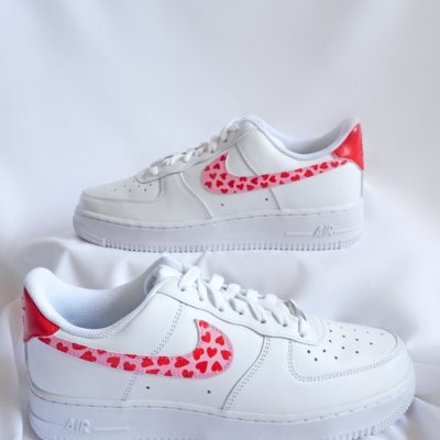 Paar weiße Nike Air Force 1 Custom Sneaker mit rotem Herz-Valentinstag-Design, viele kleine rote Herzen auf den Swoosh-Elementen, gestapelte Präsentation vor weißem Stoff