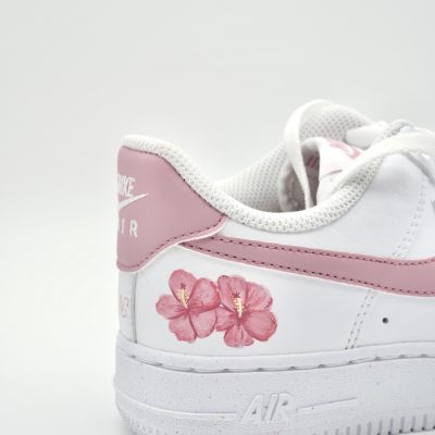 Nahaufnahme eines weißen Nike Air Force 1 mit rosa Hibiskusblüten auf der Ferse, rosa Fersenkappe und feinem rosa 'Nike Air' Schriftzug auf dem Mittelfeld.