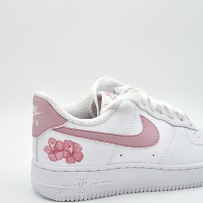 Nahaufnahme eines weißen Nike Air Force 1 mit kleiner rosa Hibiskusblüte auf dem Swoosh-Bereich, rosa Fersenkappe und feinem rosa 'Nike Air' Schriftzug an der Ferse.