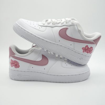 Zwei gestapelte weiße Nike Air Force 1 mit rosa Swoosh, kleinen rosa Hibiskusblüten und tropfendem rosa Akzent über dem Swoosh im frischen Sommer-Look.