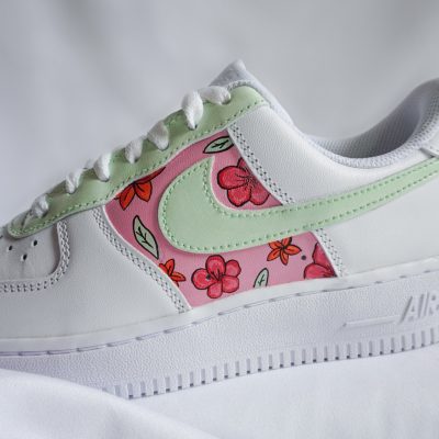 Nahaufnahme eines weißen Nike Air Force 1 mit hellgrünem Swoosh und buntem Blüten-Motiv aus rosa Hibiskusblüten und kleinen Schmetterlingen am Mittelfeld.