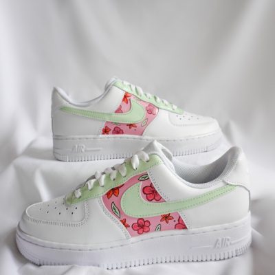 Zwei gestapelte weiße Nike Air Force 1 mit hellgrünem Swoosh und buntem Hibiskusblüten-Motiv in Rosa- und Rot-Tönen sowie kleinen Schmetterlingen am Mittelfeld.
