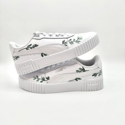 Zwei gestapelte weiße Puma Sneaker mit handgemalten grünen Eukalyptus-Zweigen entlang der Seiten im naturnahen Kommunion-Design auf hellem Hintergrund.