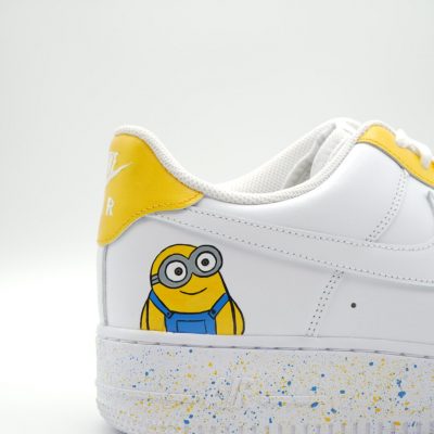 Nahaufnahme eines weißen Nike Air Force 1 mit handgemaltem fröhlichen Minion in blauer Latzhose, gelber Fersenkappe und blau-gelben Farbspritzern auf der Sohle.