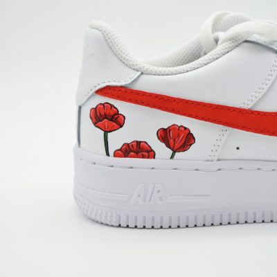 Nahaufnahme eines weißen Nike Air Force 1 mit rotem Swoosh und kleinen handgemalten roten Mohnblumen unter dem Swoosh als zartes florales Detail.