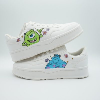 Zwei gestapelte weiße Plateau-Sneaker mit handgemalten Monster AG Charakteren Mike und Sulley sowie verteilten lila Blumen und Sternchen auf den Seiten.