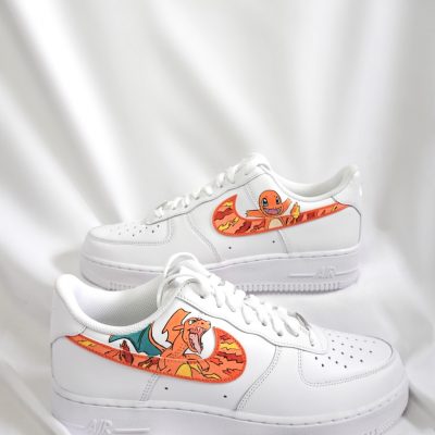 Paar weiße Nike Air Force 1 Custom Sneaker mit Pokémon Glumanda Design, orange-gelbe Flammen-Akzente auf dem Mittelfußbereich, gestapelte Präsentation vor weißem Stoff