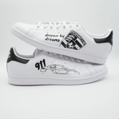 Zwei weiße Adidas Stan Smith mit handgemalter Porsche 911 Silhouette, Porsche-Logo, schwarzer Fersenkappe und Aufschrift 'driven by dreams' am Mittelfeld.