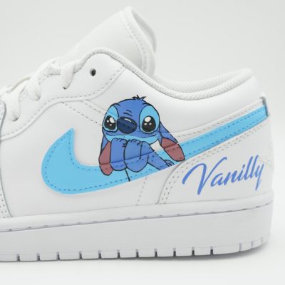 Nahaufnahme eines weißen Air Jordan 1 Low mit blauem Stitch aus Lilo & Stitch hängend am Swoosh und blauem Schriftzug 'Vanilly' an der Ferse.