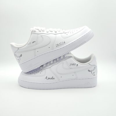 Zwei gestapelte weiße Nike Air Force 1 Taufsneaker mit feinen Storch-Silhouetten, Datum '28.12.24', Geburtsgewicht '2980 g' und Namen 'Amelie' in feiner Linie.