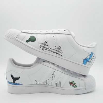 Paar weiße Adidas Superstar Custom Sneaker mit Reise-Motiven, Golden Gate Bridge Heißluftballon Walflosse und New York Skyline als Linienzeichnungen