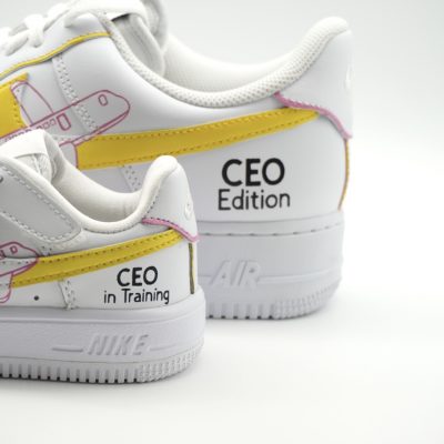 Nahaufnahme zweier weißer Nike Air Force 1 Sneaker im 'CEO Edition' und 'CEO in Training' Design mit gelbem Swoosh, lila Linien und kleinen Flugzeug-Motiven.