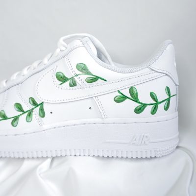 Ein weißer Sneaker mit handgemaltem, grünem Blätterranken-Motiv entlang der Seitennaht und des Logos. Der Schuh liegt auf einem weißen Stoffhintergrund.
